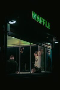 Moody night scene of a waffle shop front with glowing neon sign and interior lighting.