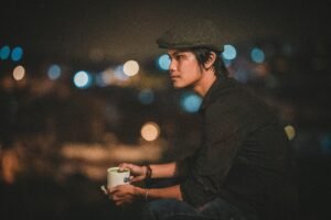 Young man in a cap enjoying coffee outdoors at night with city bokeh lights.